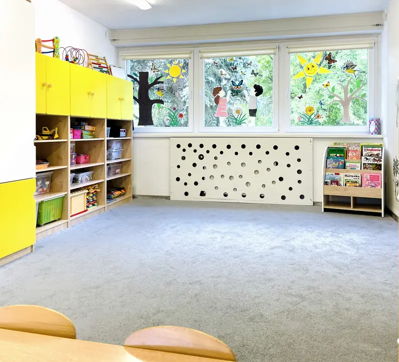 Słoneczna sala z dużymi oknami na ogród, żółtymi szafkami i półkami z zabawkami