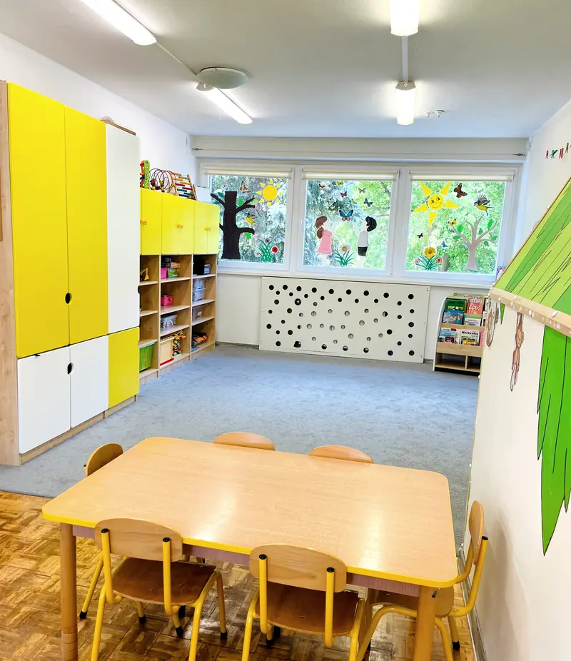 Sala zajęć ze stolikiem i krzesełkami, żółtymi szafkami i oknami na ogród