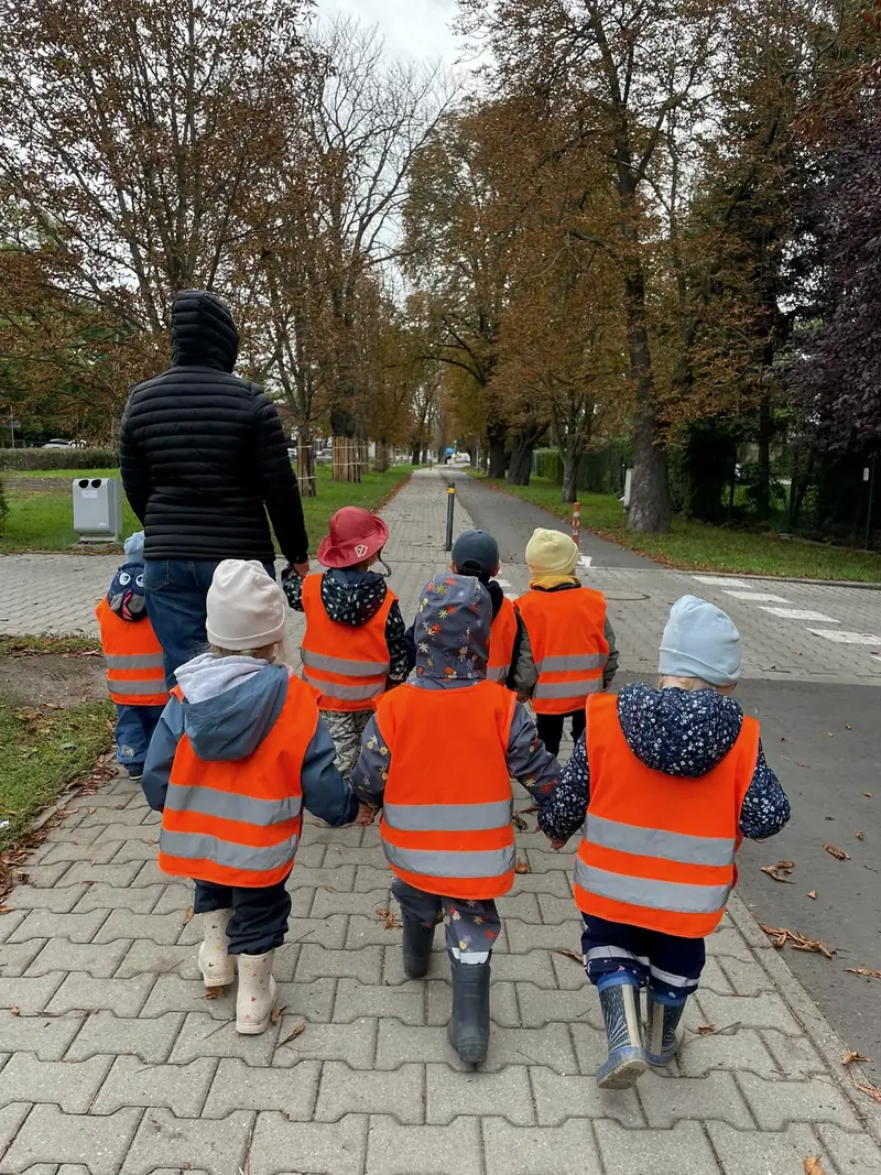 Dzieci w odblaskowych kamizelkach idą z opiekunką na spacer aleją drzew